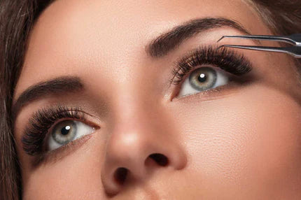 A person is using tweezers to apply false eyelashes, focusing on the eye area with long, voluminous lashes.